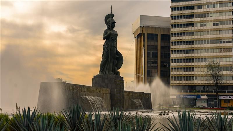 Estatua en una fuente al atardecer, junto a edificios modernos, con agua salpicando a su alrededor.