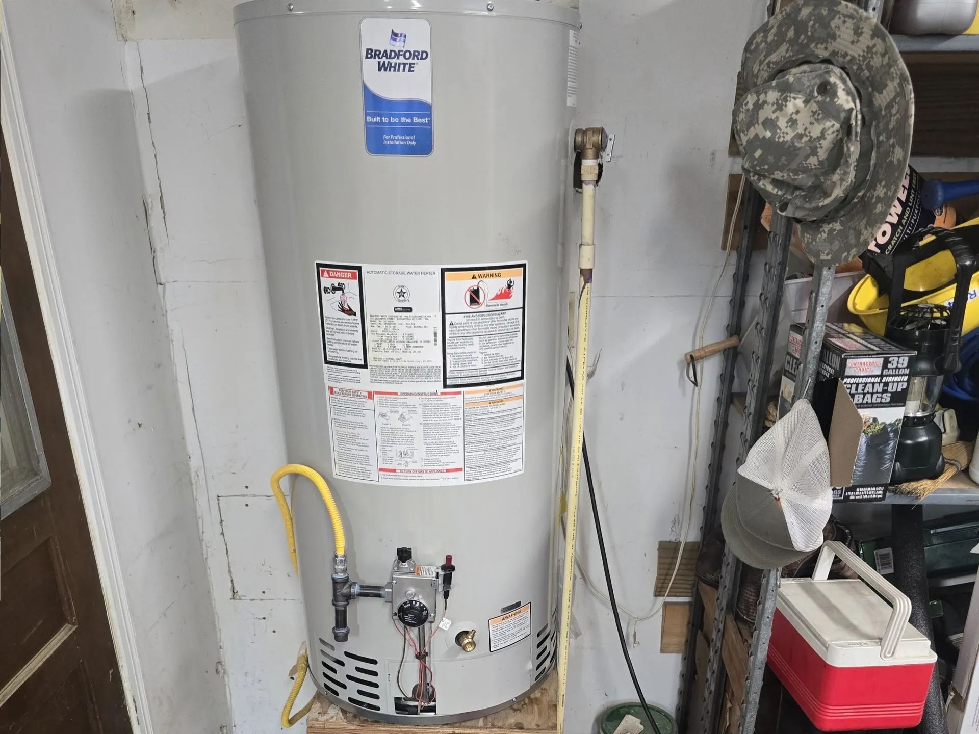 Water heater in a utility room, with pipes and warning labels, beside a vacuum and storage items.