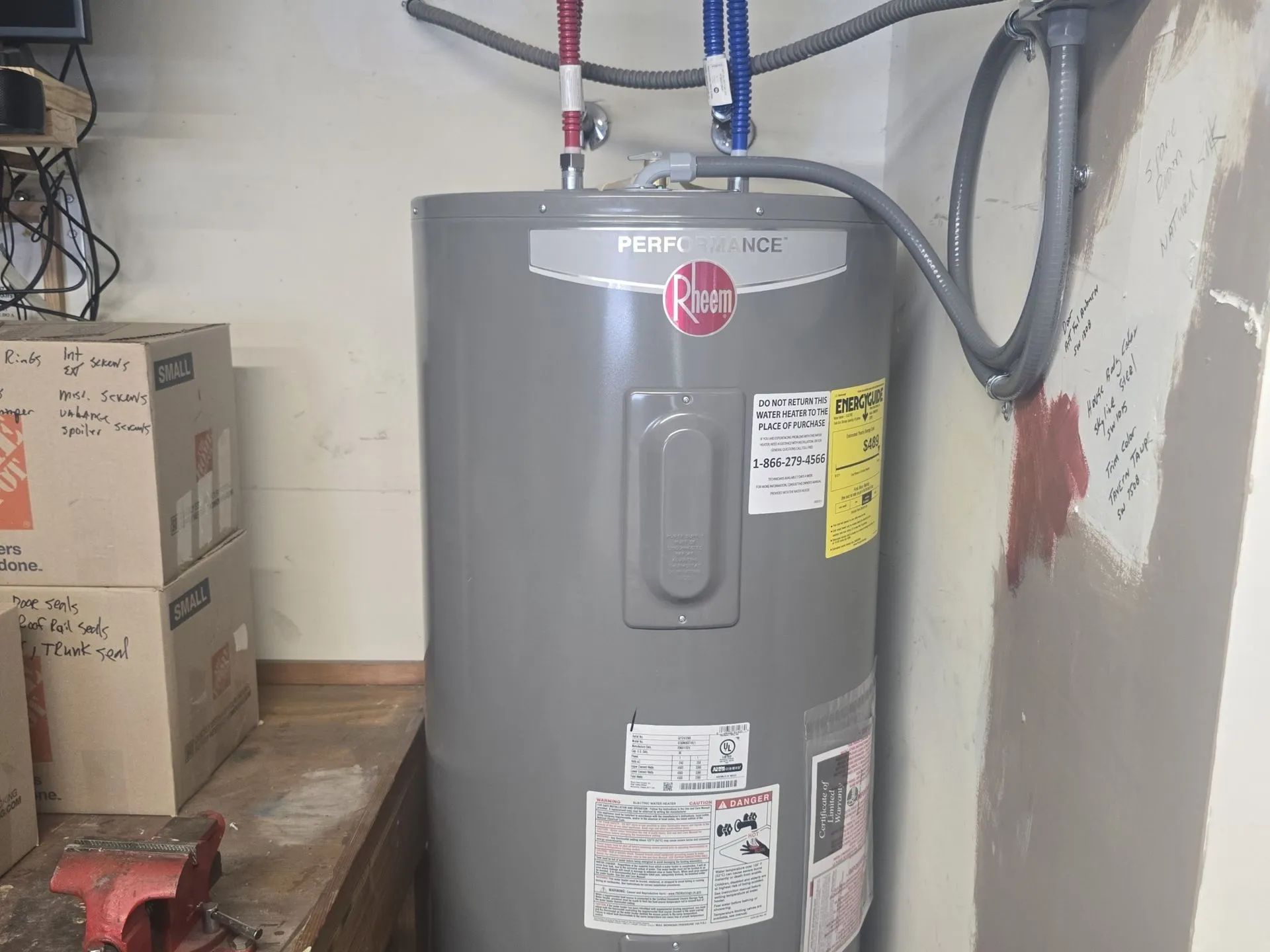 Gray Rheem water heater in a utility room with stacked boxes and exposed pipes.