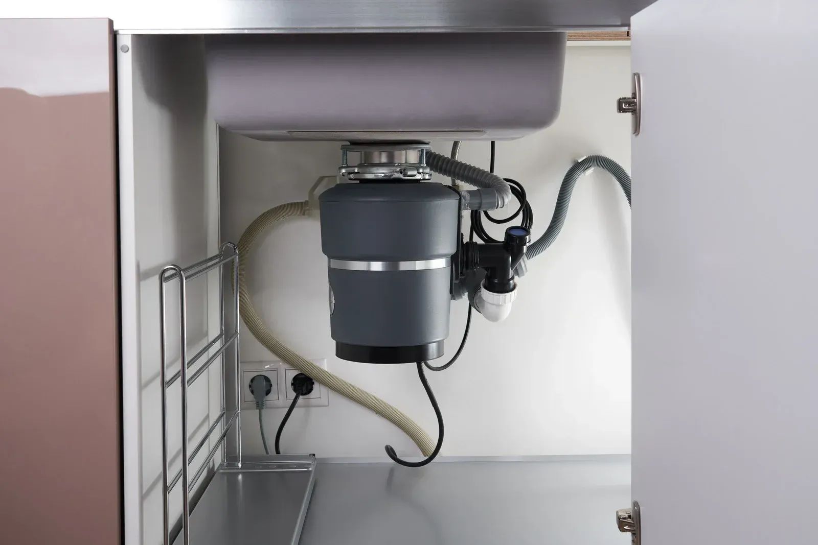 Under-sink view of a gray garbage disposal with plumbing and hoses inside a cabinet