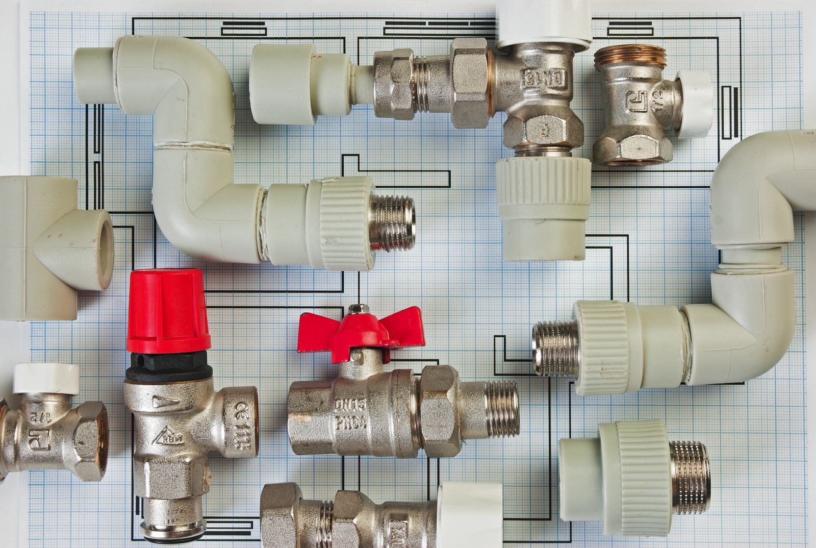 Plumbing pipe fittings and valves arranged on graph paper, including white PVC and red-handled metal parts