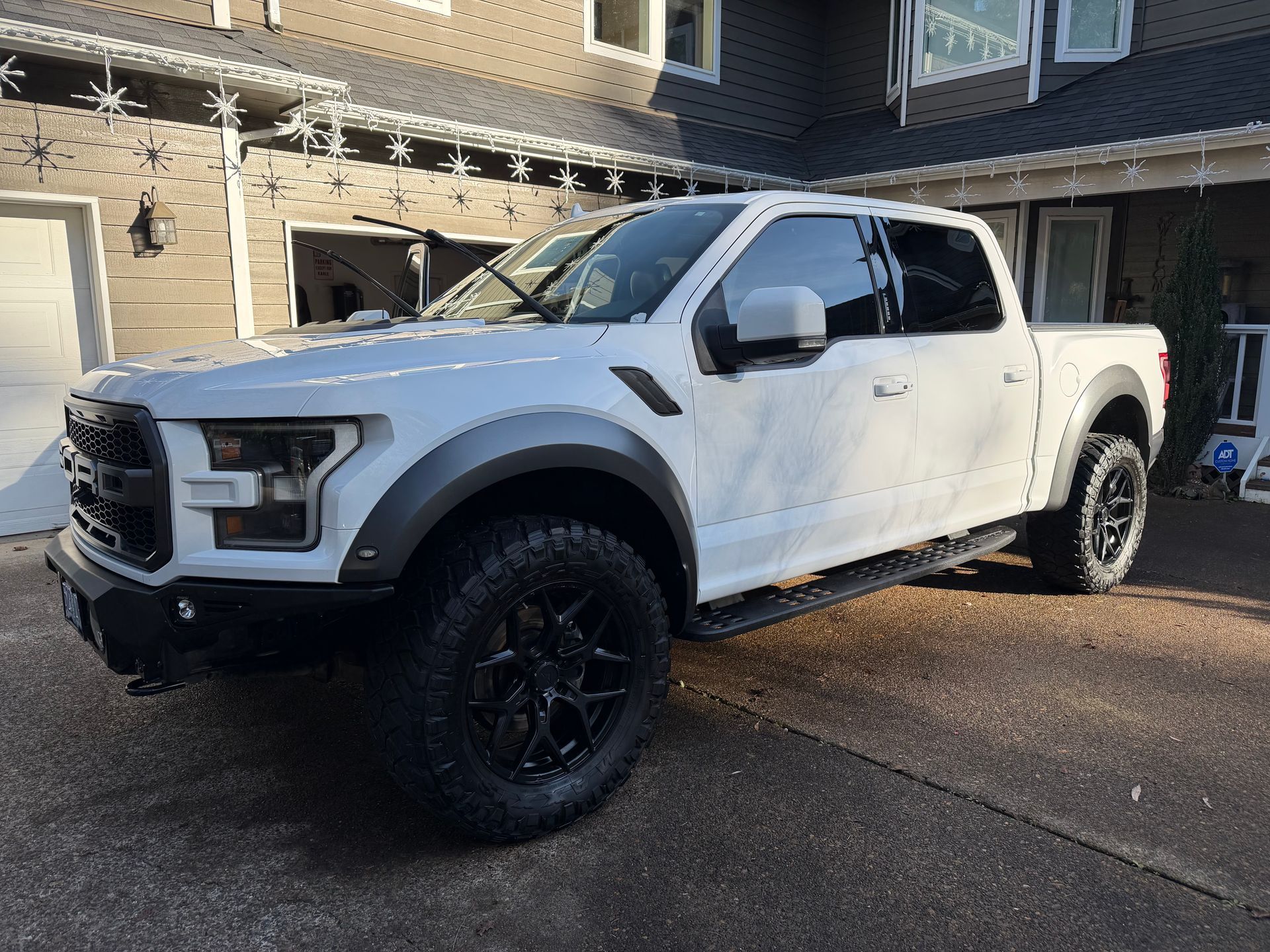 White Ford Raptor truck parked in front of a house, black wheels and trim, overcast lighting.