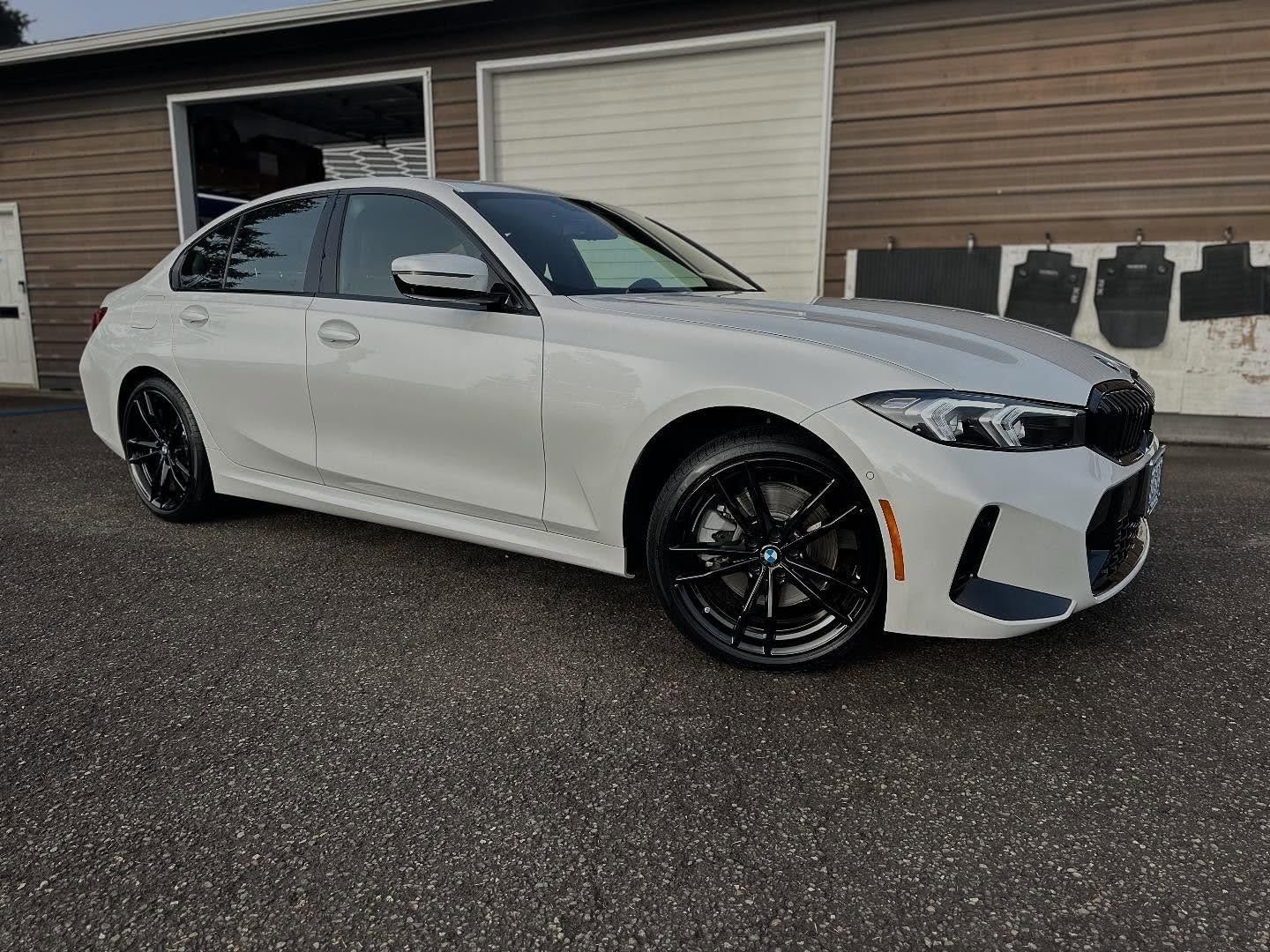 A white BMW 3 Series sedan parked on asphalt in front of a building with large garage doors.