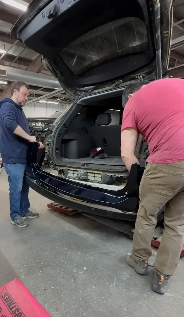 Two people working in a garage, installing a dark car bumper onto the rear of a vehicle with an open trunk.