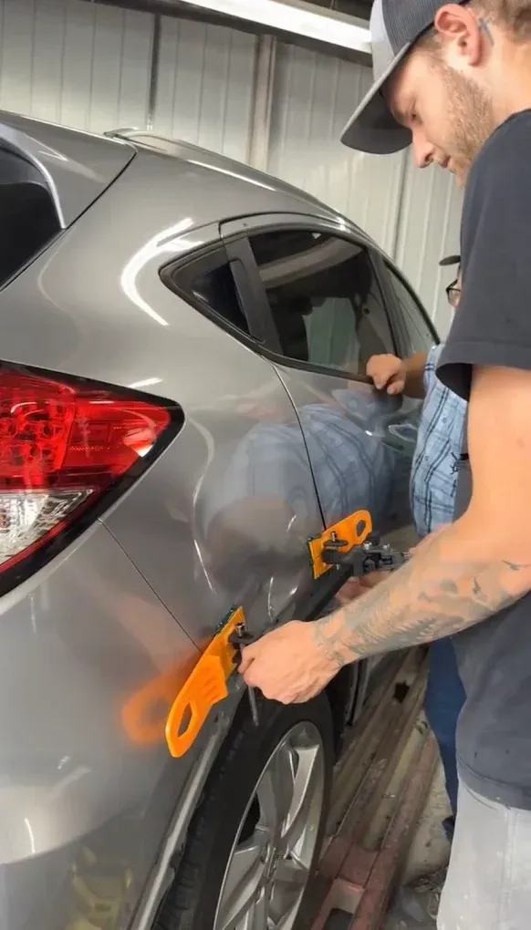 A person uses orange dent repair tools on the rear quarter panel of a silver car in a workshop.