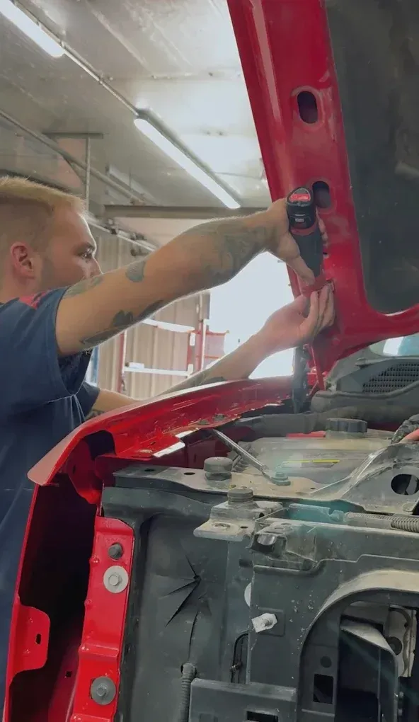 A mechanic working on the hood hinge of a red truck in an auto repair shop.