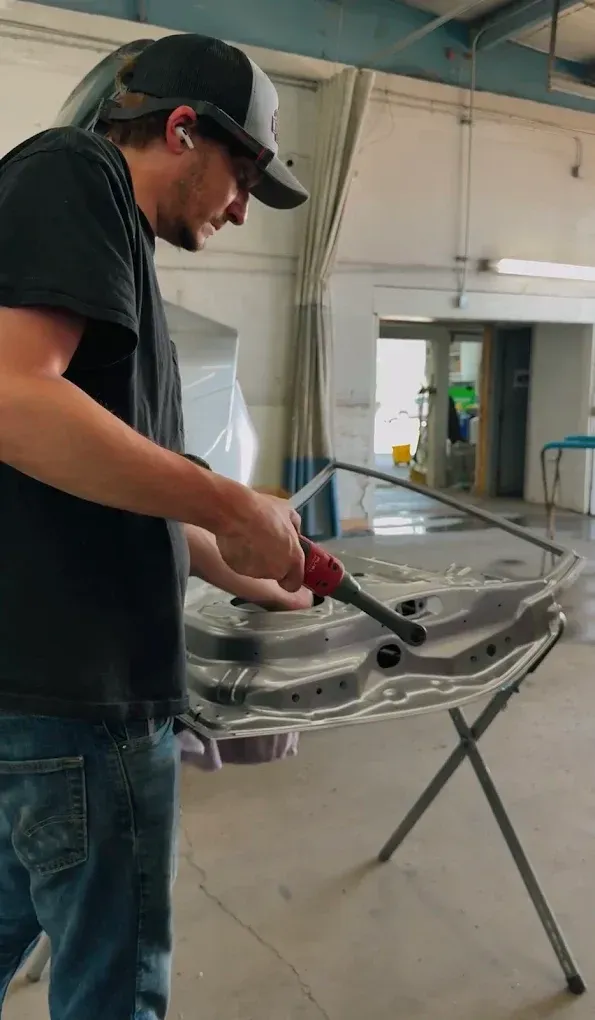 A person in an auto body shop uses a handheld power tool to sand the inner frame of a car door mounted on a metal stand.
