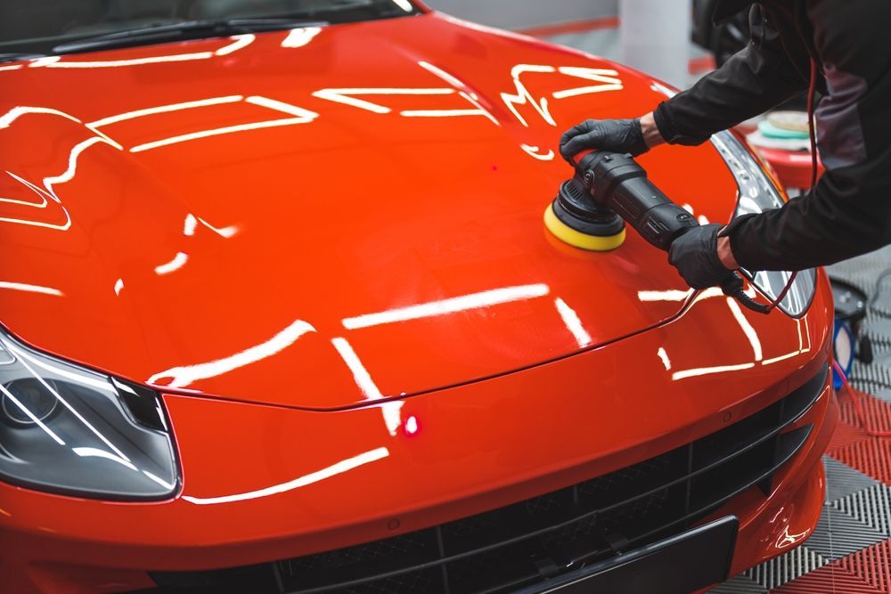 A technician using a dual-action polisher to buff the bright orange hood of a luxury sports car in a workshop.