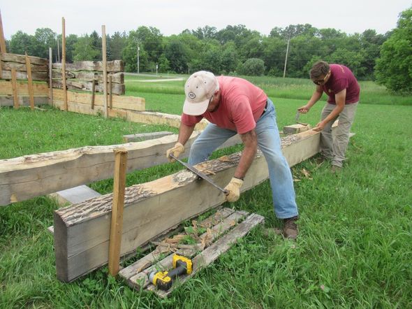 Two people sawing a large wooden beam outdoors. One is using a hand saw, another holds the beam steady.