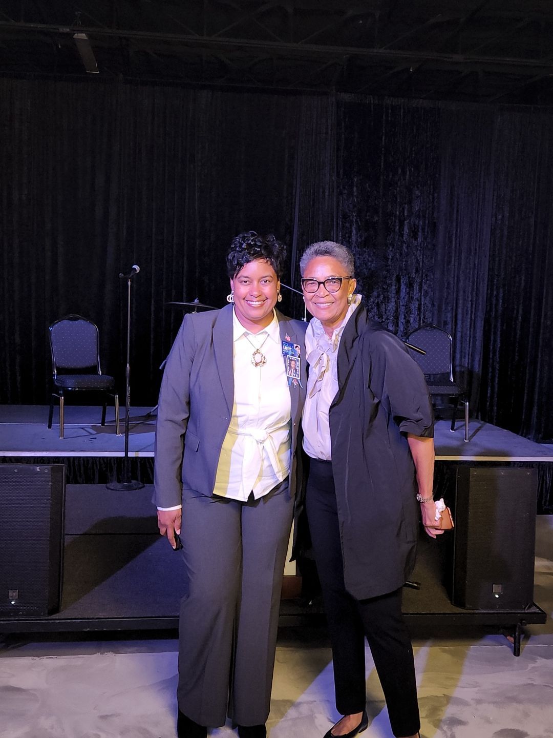 Two people smiling and posing on a dimly lit stage in front of black curtains.