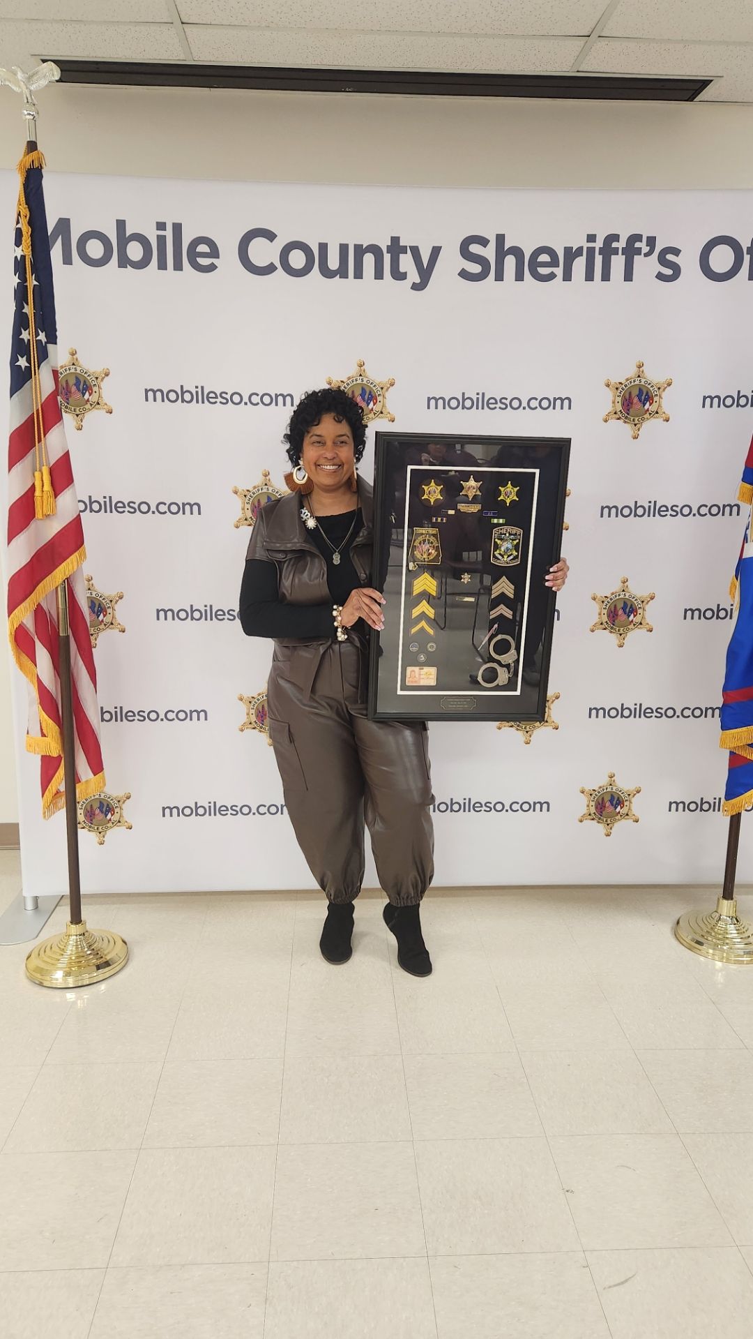 Woman holding framed award beside sheriff’s office backdrop and U.S. flag