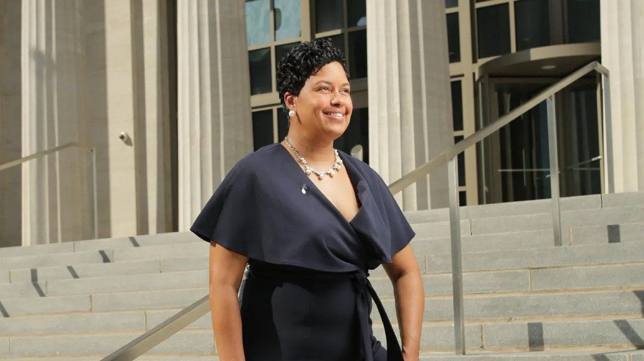 Woman in a dark dress smiling outside a building with steps and glass doors