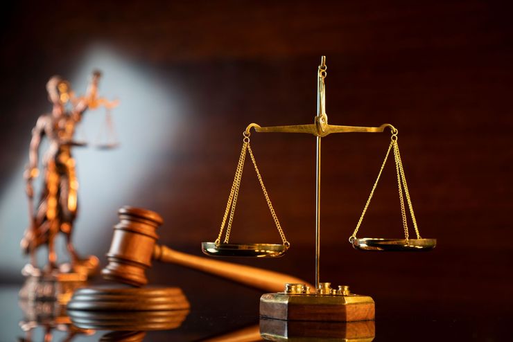Gold justice scales and gavel on a dark wooden background, with a blurred Lady Justice statue.