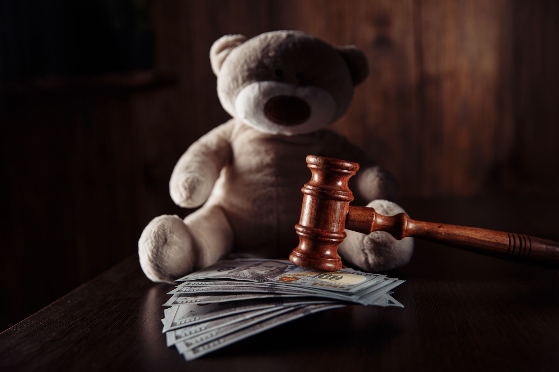 Teddy bear beside a gavel and a stack of money on a dark table, suggesting a legal or auction setting