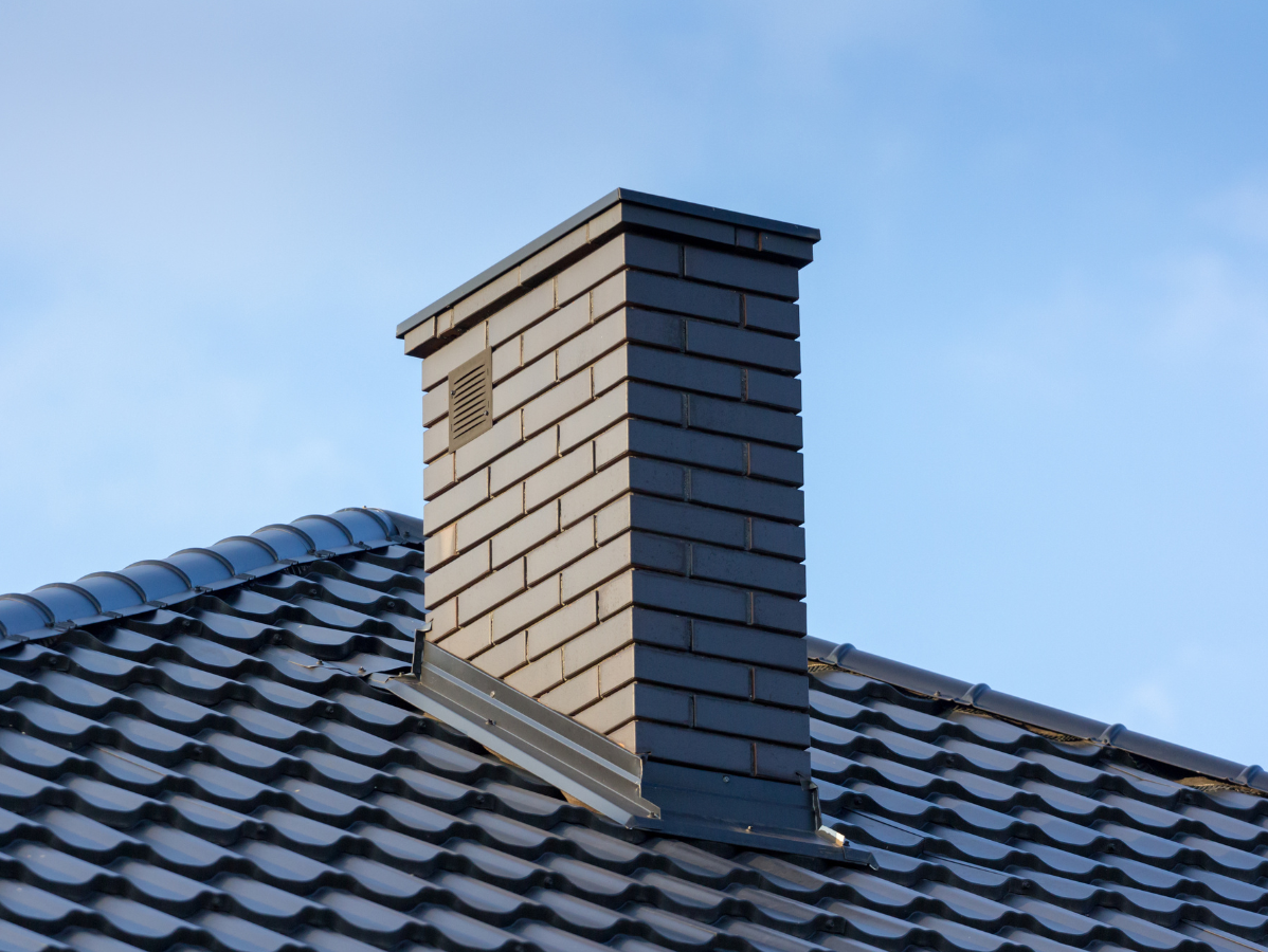 A gray brick chimney sits on a dark-tiled roof against a clear blue sky.