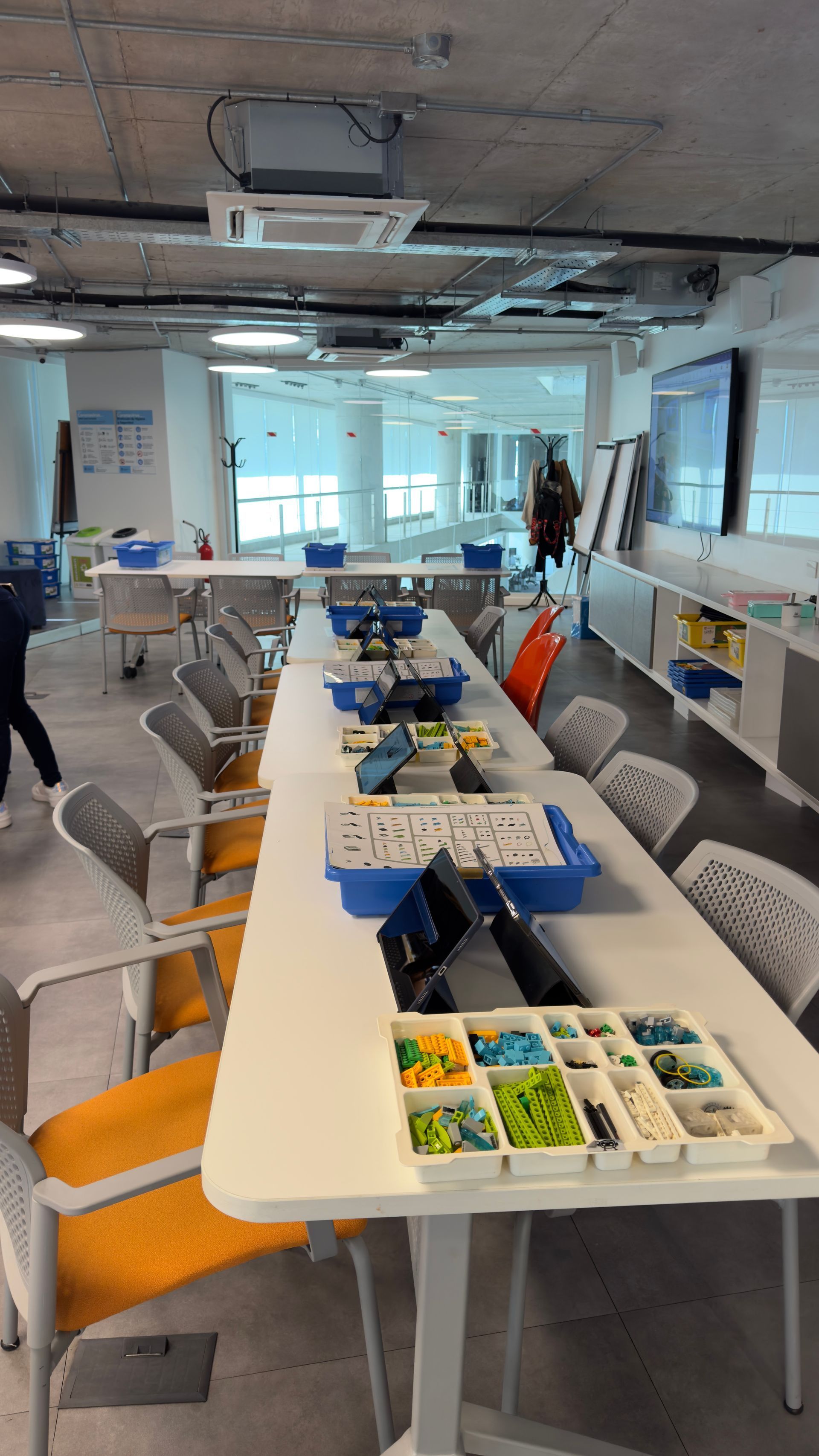 Sala de reuniones con una mesa blanca larga, sillas y coloridas bandejas de materiales en un espacio de oficina luminoso.