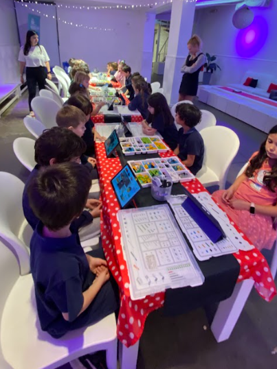 Niños sentados en una mesa larga con ordenadores portátiles, jugando a un juego en una sala de fiestas muy iluminada.