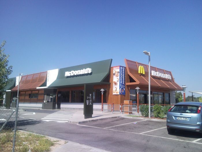 Exterior de un restaurante McDonald's con carril de autoservicio y un coche aparcado bajo un cielo azul despejado.