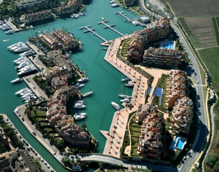 Vista aérea de un puerto deportivo con yates, canales y edificios de color beige a orillas del agua.