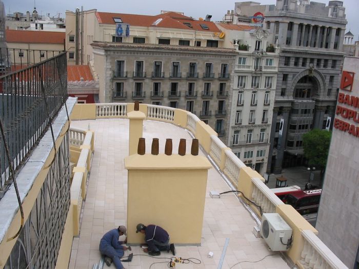 Obreros reparando la terraza de una azotea cerca de edificios de la ciudad, con herramientas y cables en el suelo.