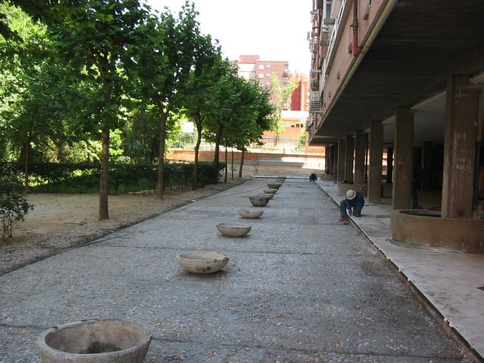 Paseo al aire libre con bancos circulares de hormigón dispersos junto a árboles y un edificio bajo un alero cubierto.