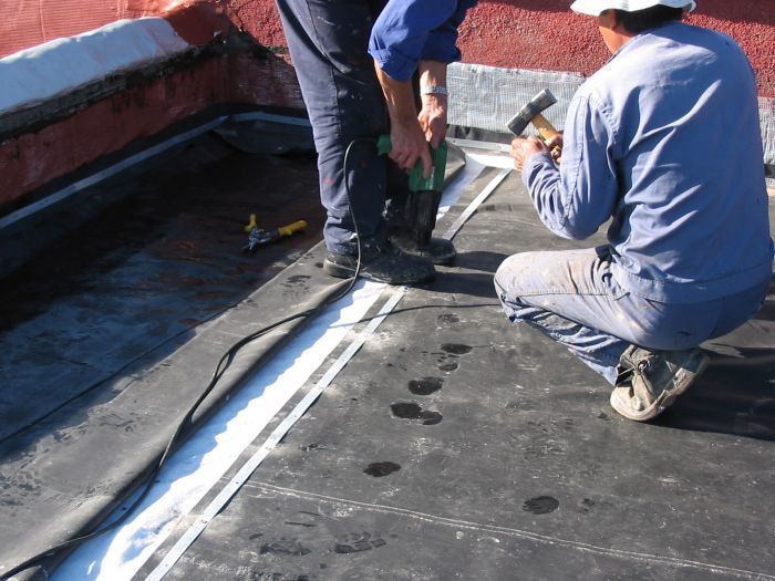 Dos trabajadores reparando una junta del tejado con material impermeabilizante negro y herramientas.