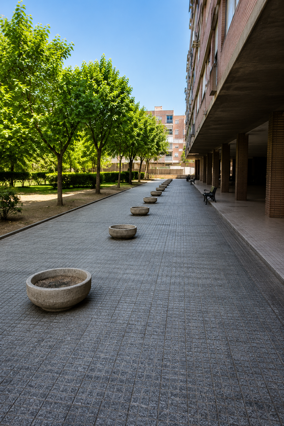 Pasarela exterior con jardineras de piedra junto a un edificio y árboles en un patio.