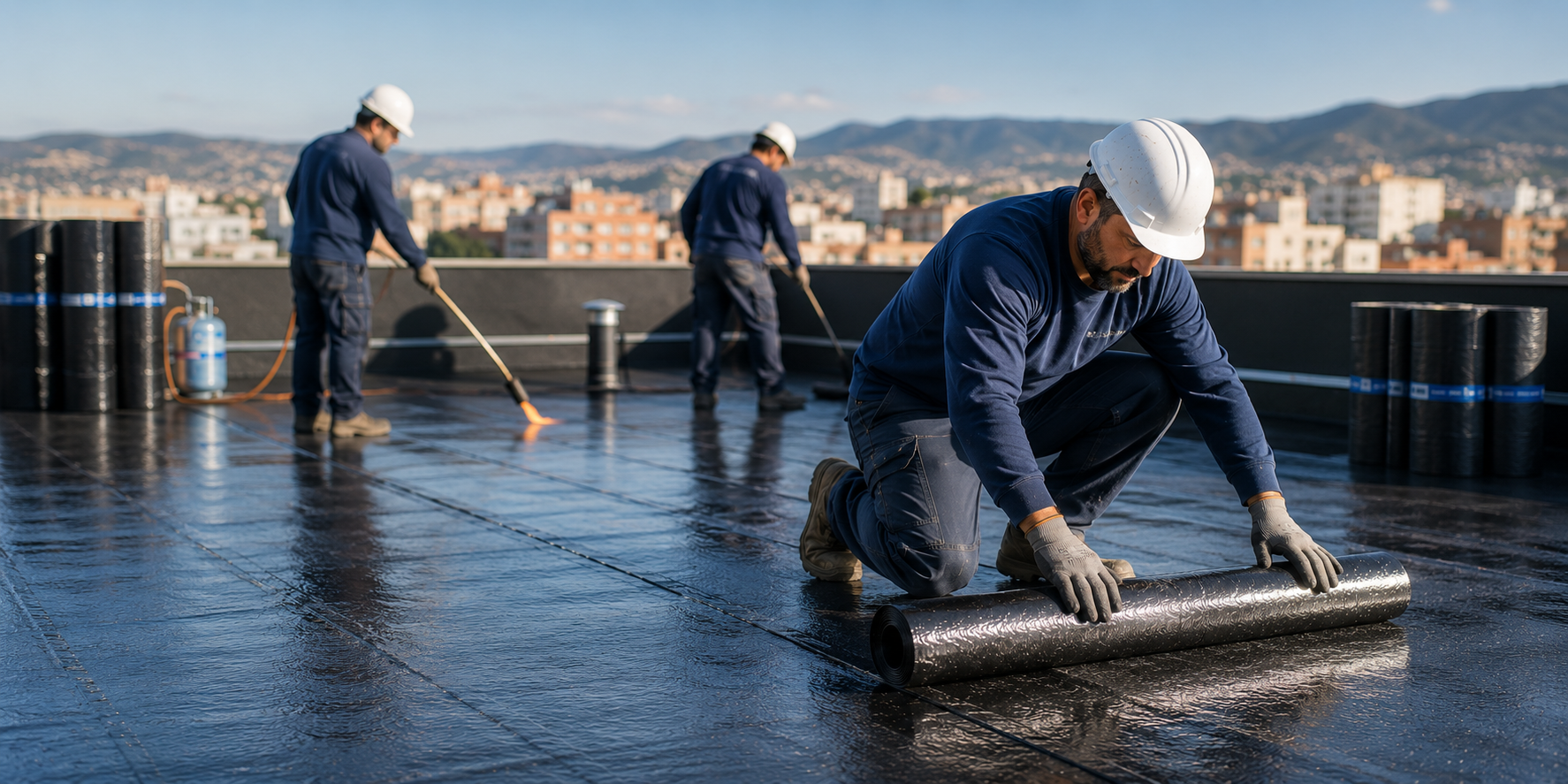 Trabajadores colocando material de techado negro en un techo plano con edificios de la ciudad y montañas al fondo.