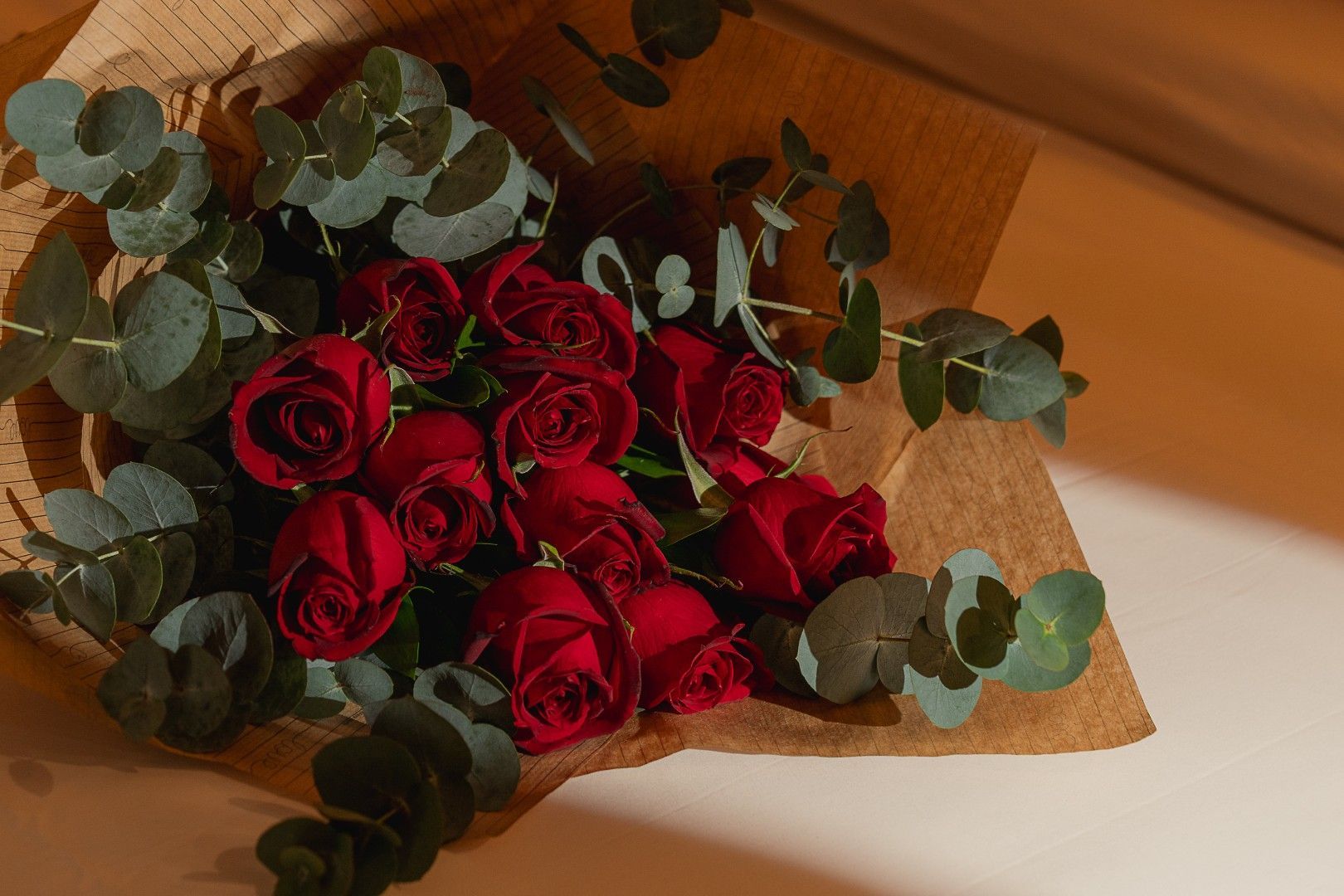 Um buquê de rosas vermelhas escuras envolto em papel kraft rústico e decorado com raminhos de eucalipto sobre um fundo neutro.