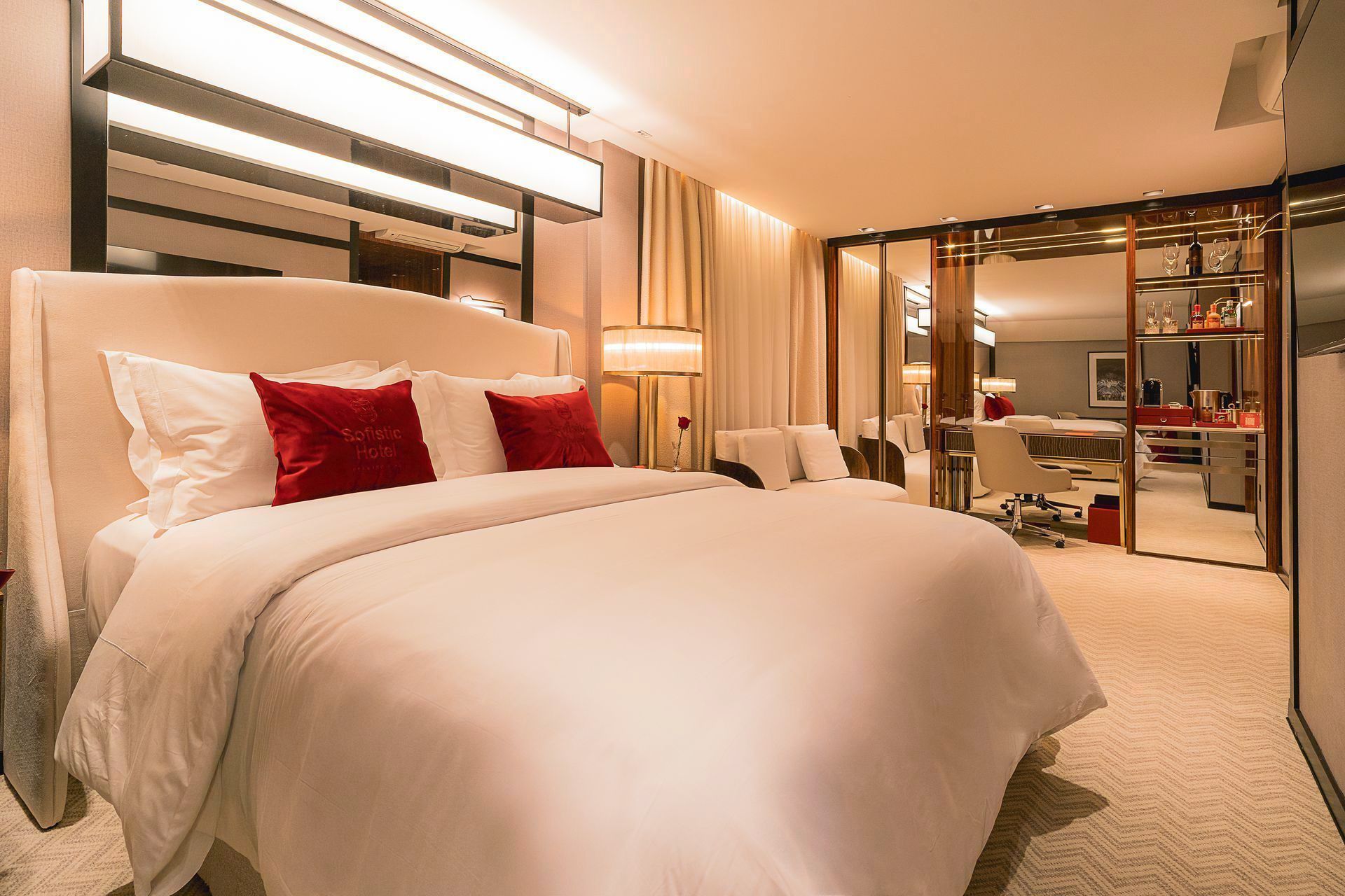 Um quarto de hotel luxuoso e bem iluminado, com uma cama macia de lençóis brancos e almofadas decorativas vermelhas, ao lado de uma sala de estar com paredes de vidro.