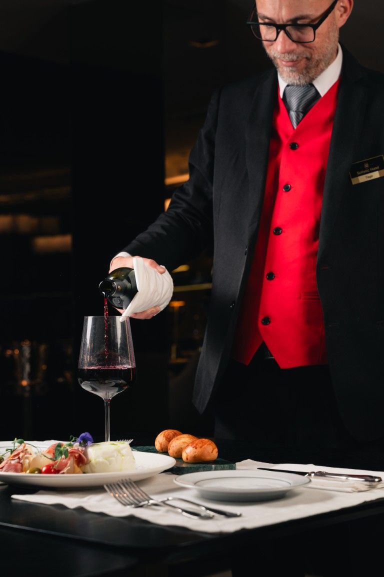Um garçom de terno preto e colete vermelho serve vinho tinto em uma taça em uma mesa posta com jantar e pãezinhos.