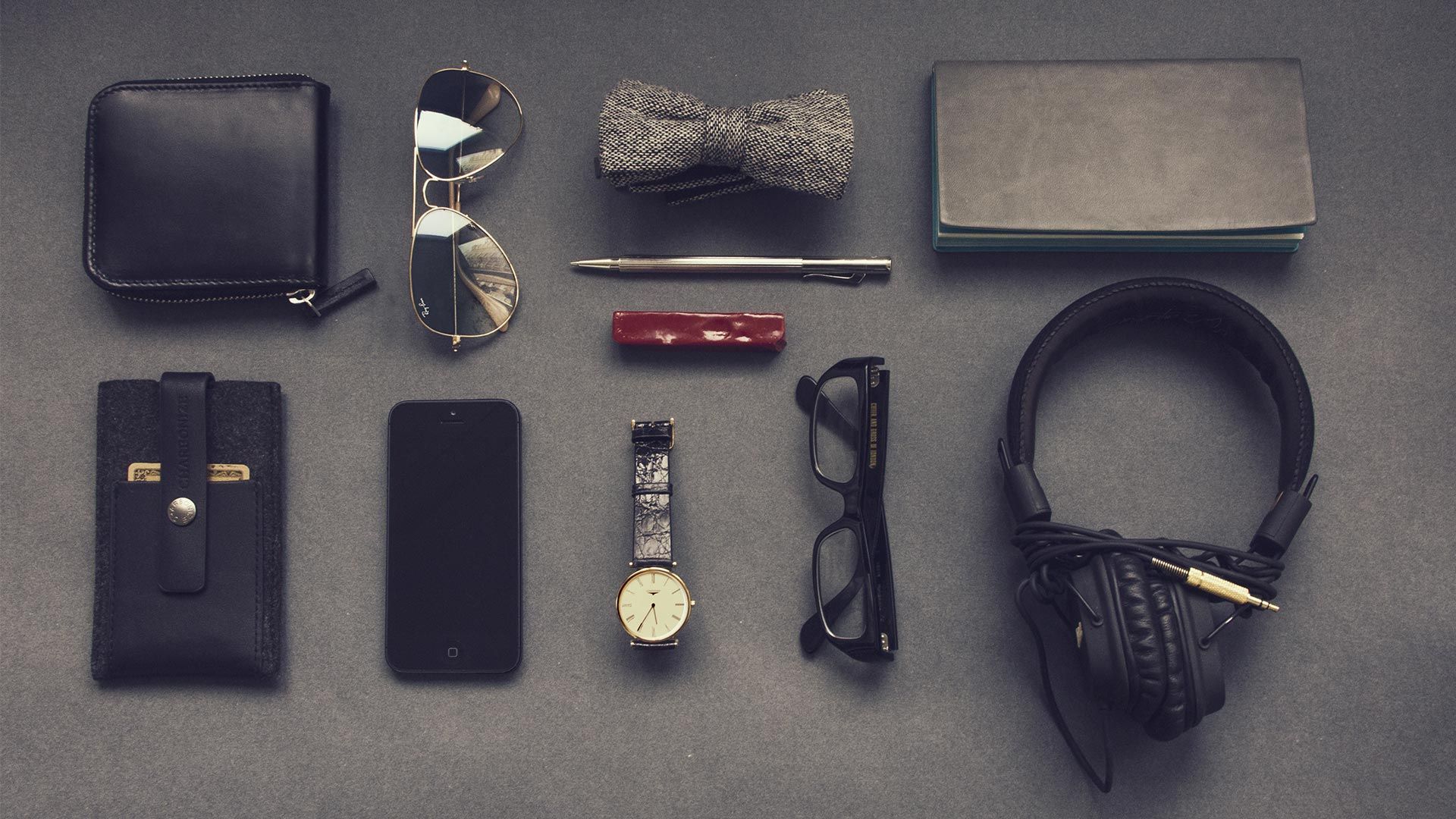 Flat lay of men's accessories: wallet, sunglasses, bowtie, notepad, pen, flashlight, phone, watch, glasses, and headphones.