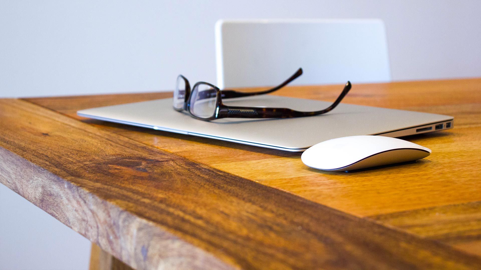 Laptop, glasses, and mouse on a wooden desk, with a white chair in the background.
