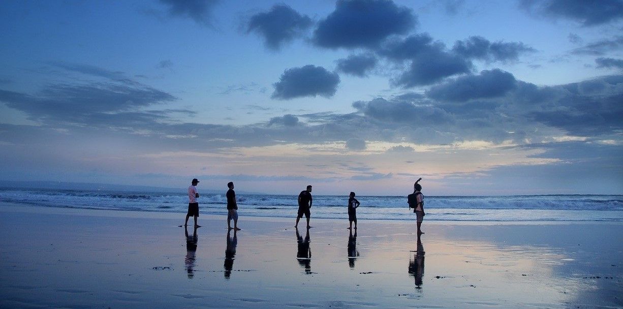 People walking on a beach at dusk, reflecting in the wet sand with a blue sky.