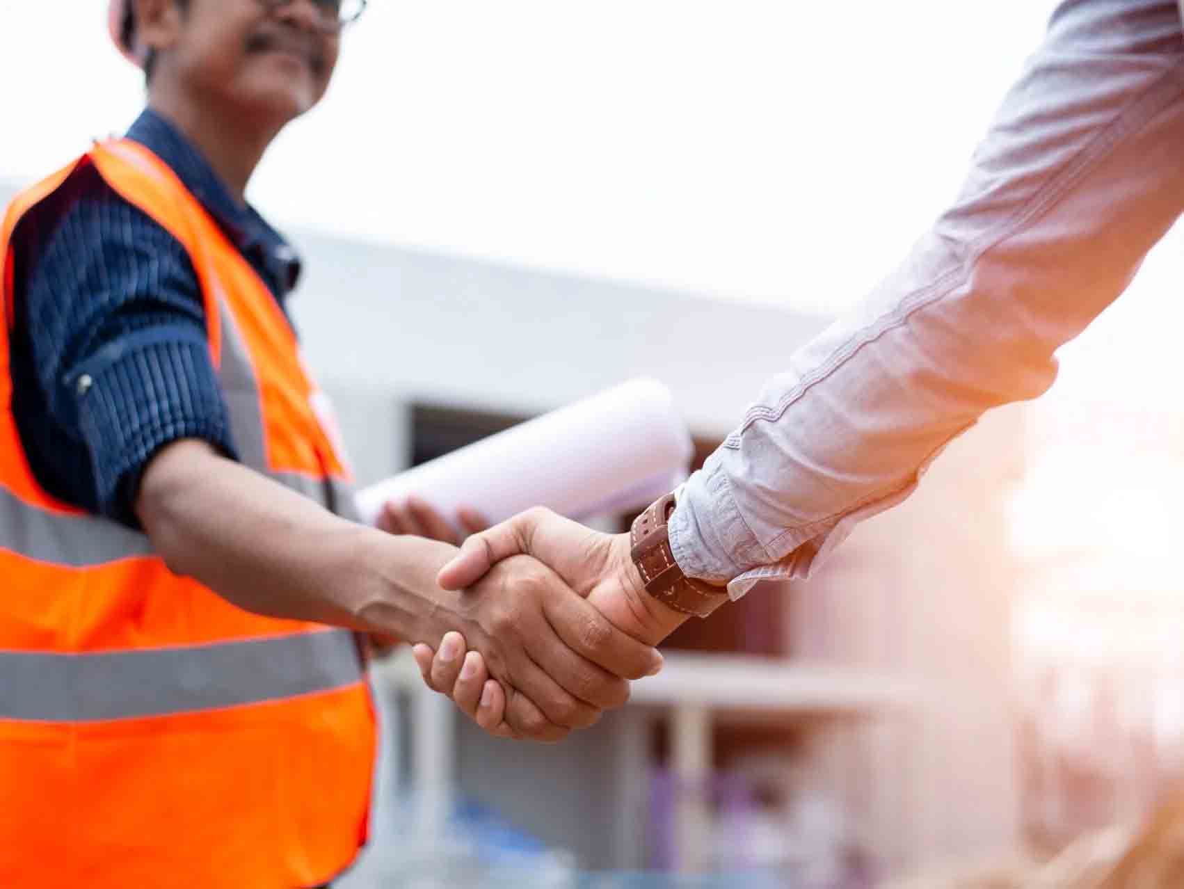 Un ouvrier du bâtiment serre la main d'une autre personne, tous deux portant des gilets de sécurité, sur un chantier.