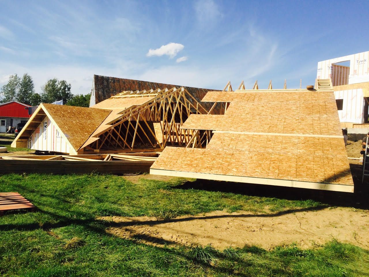 Structure en bois partiellement construite avec des toits en pente, située sur un terrain herbeux en plein air sous un ciel bleu.