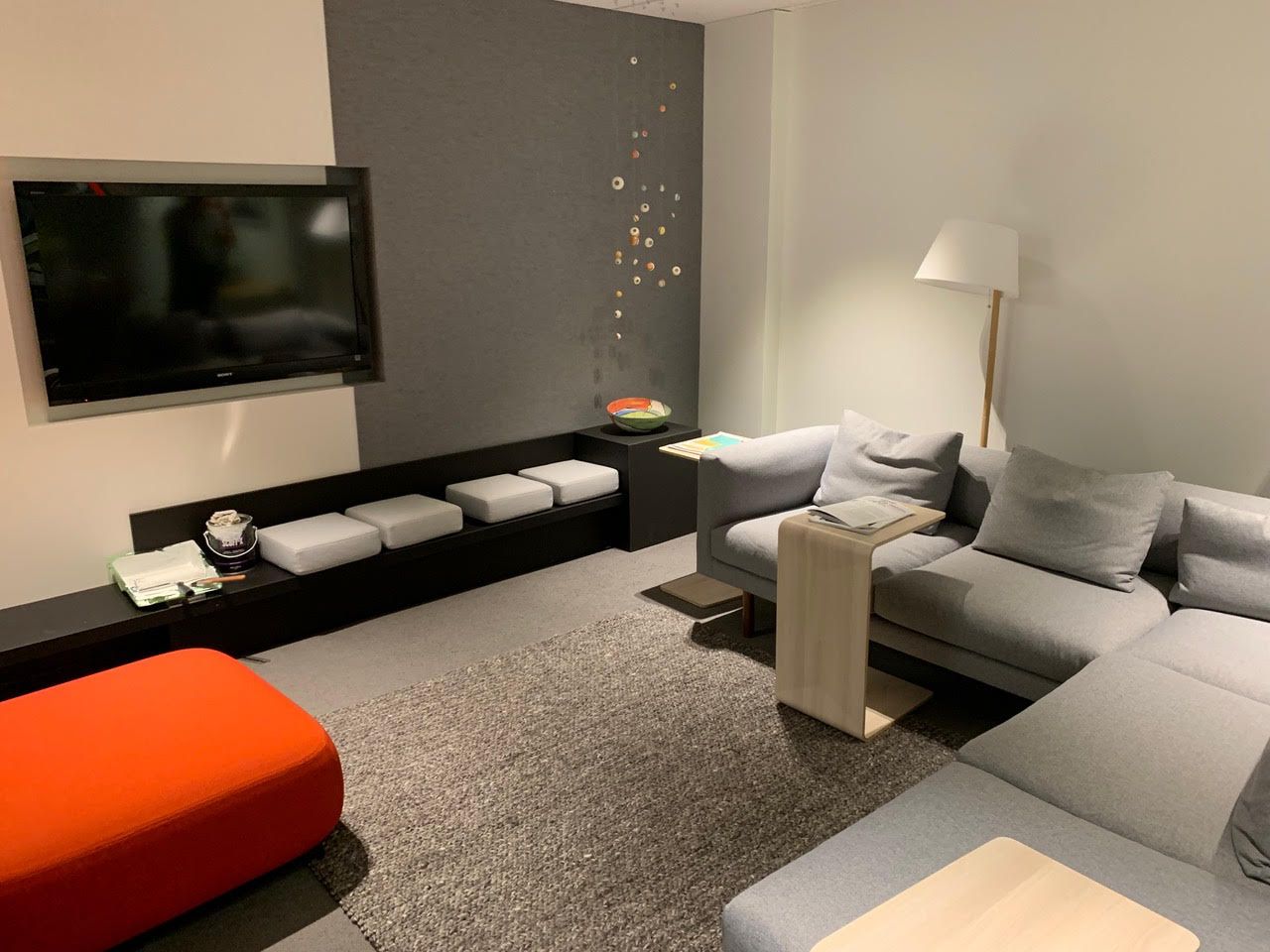 Salon moderne avec canapé d'angle gris, télévision murale, pouf orange et lampadaire