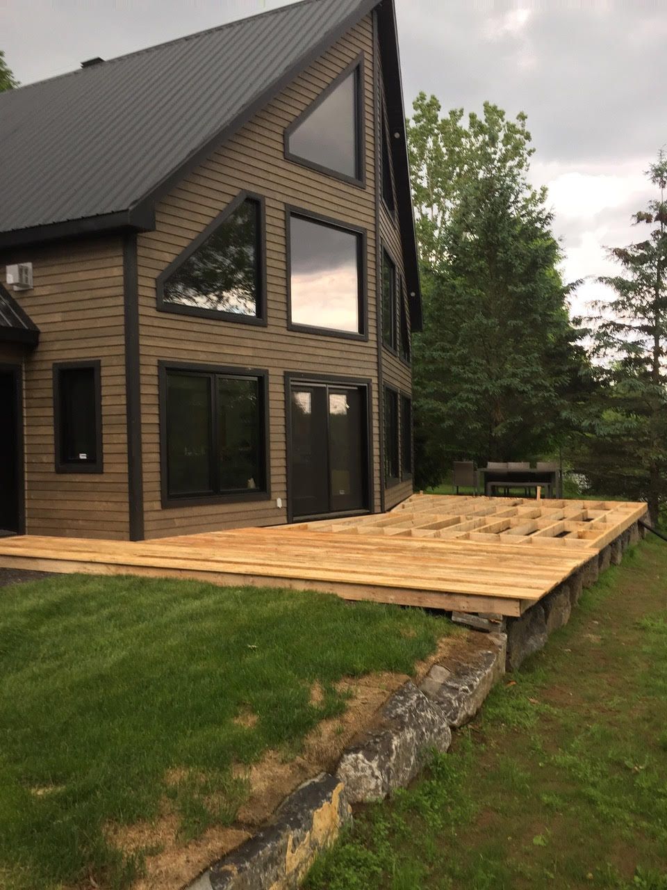 Maison moderne noire avec de grandes fenêtres et une terrasse en bois donnant sur un jardin gazonné et arboré.