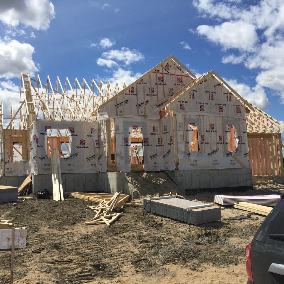 Maison partiellement construite avec pare-pluie blanc, charpente de toit apparente et matériaux de construction sur terrain en terre battue