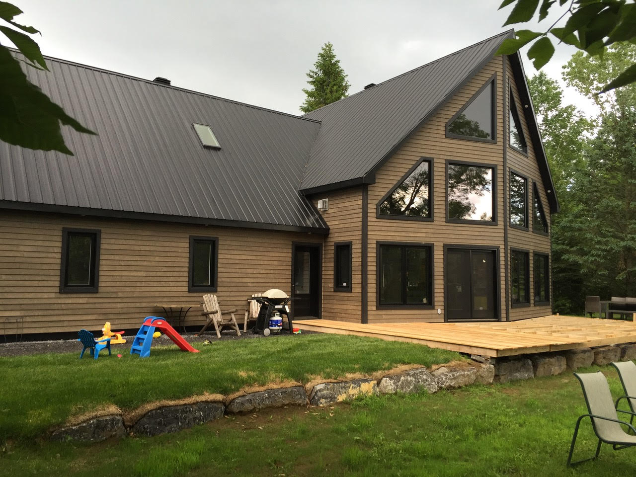Maison moderne en bois avec toit foncé et grandes fenêtres, donnant sur une pelouse avec chaises et jouets pour enfants