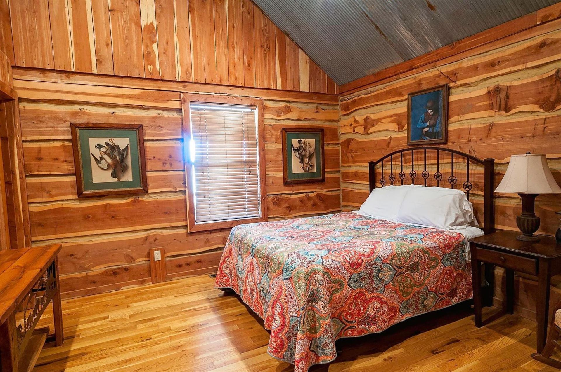 Bedroom with wooden walls, bed, window with blinds, artwork, and lamp.