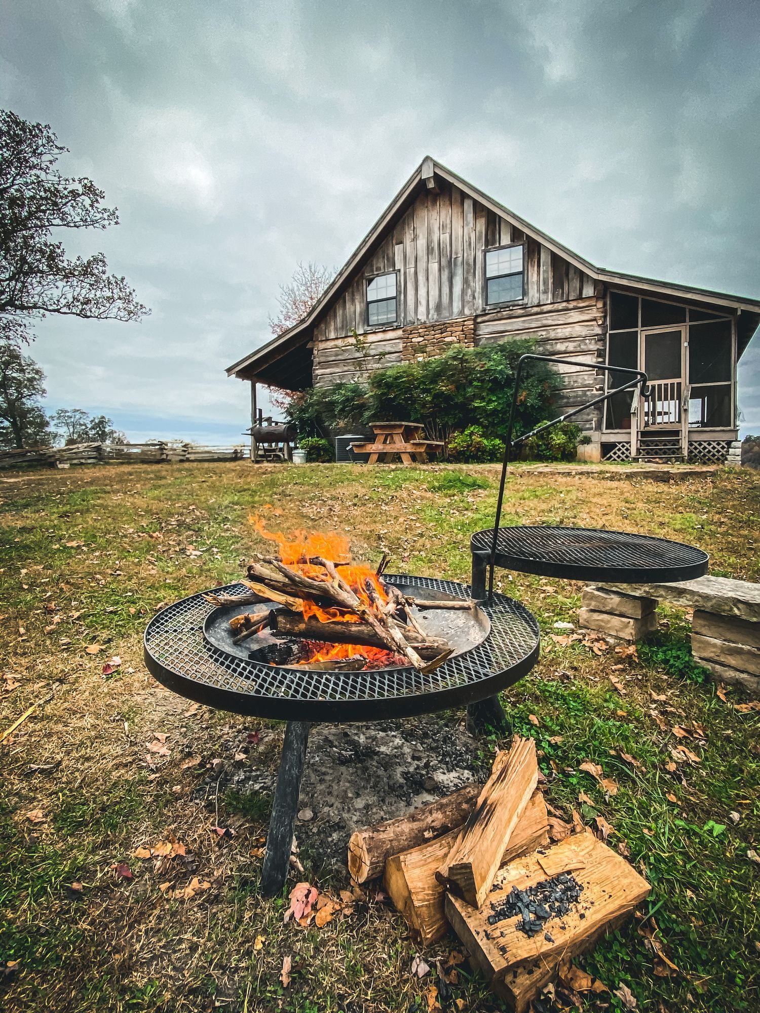 Fire pit with burning logs in front of a rustic wooden cabin on a cloudy day.