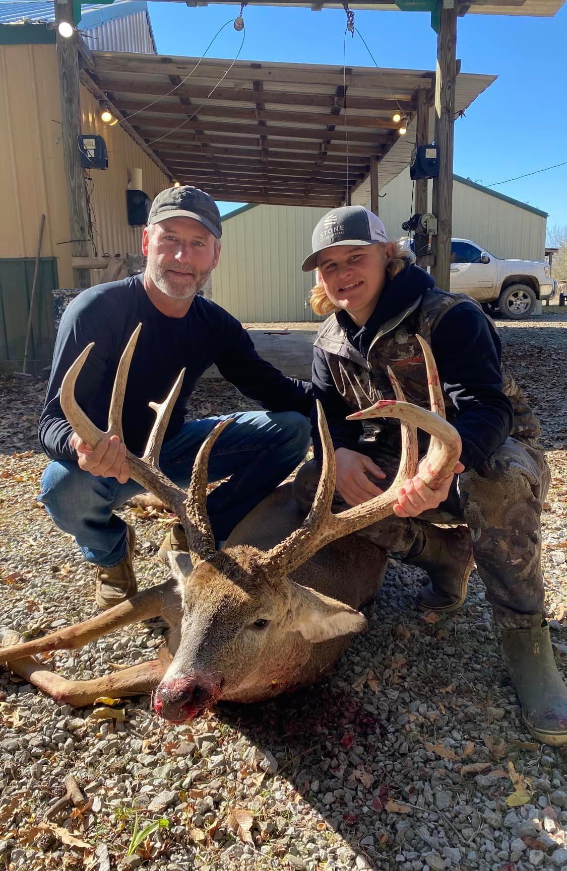 Two hunters kneel beside a large deer with antlers outside a rustic building.