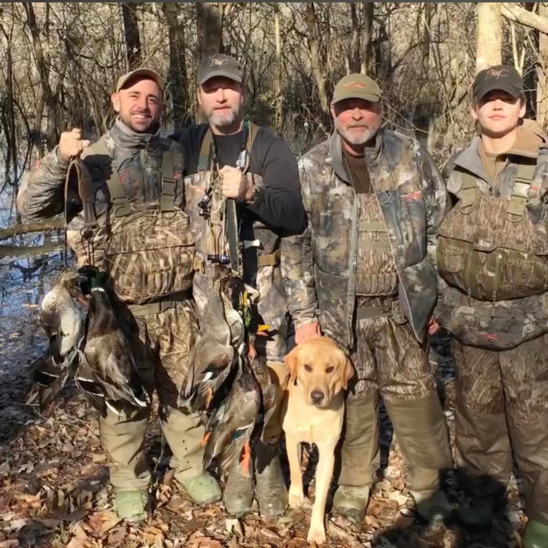 Four hunters in camouflage stand with a dog in a leafless wooded area, holding hunting gear.