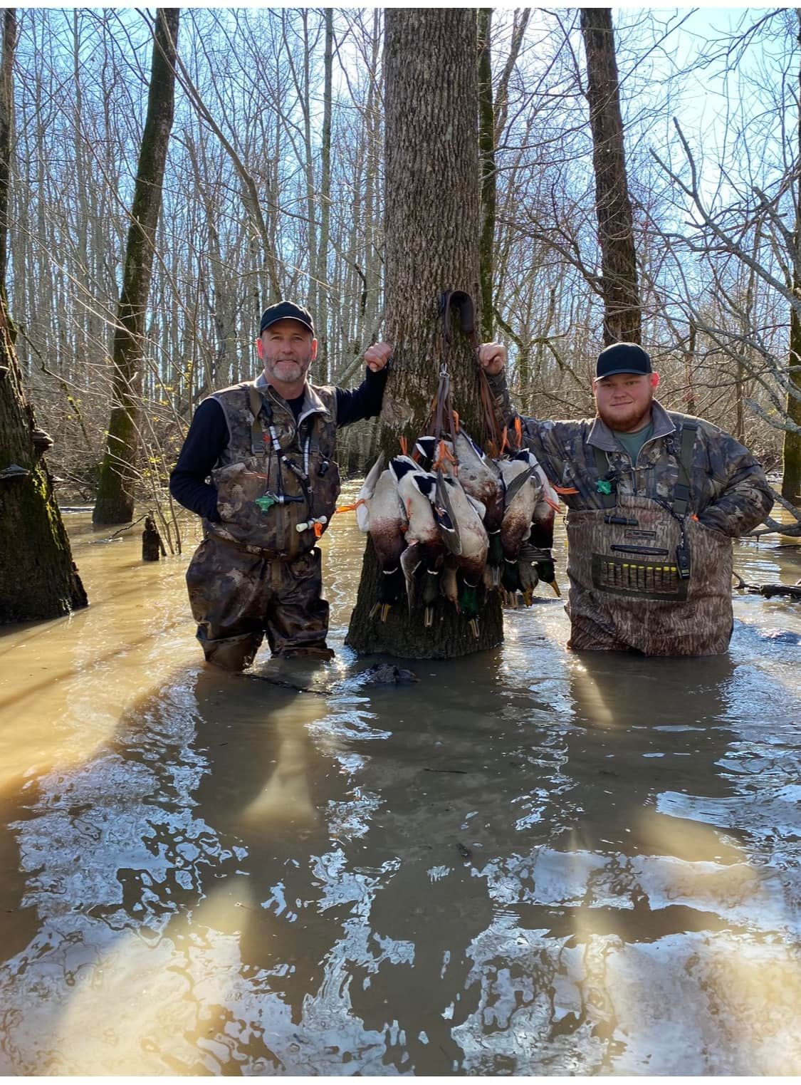 Three hunters kneel in flooded woods holding a display of harvested ducks.