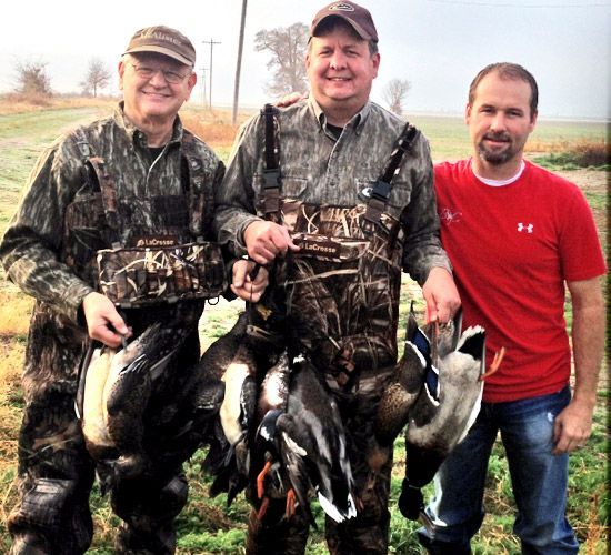 Three men outdoors holding several dead ducks and geese after a hunt