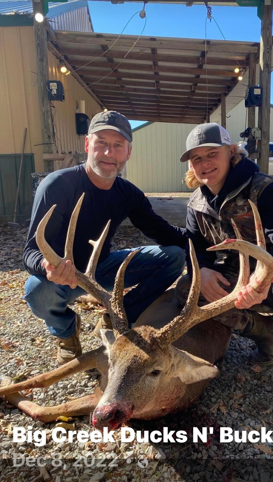 Two people kneel beside a large buck with a giant antler rack under a wooden shelter.