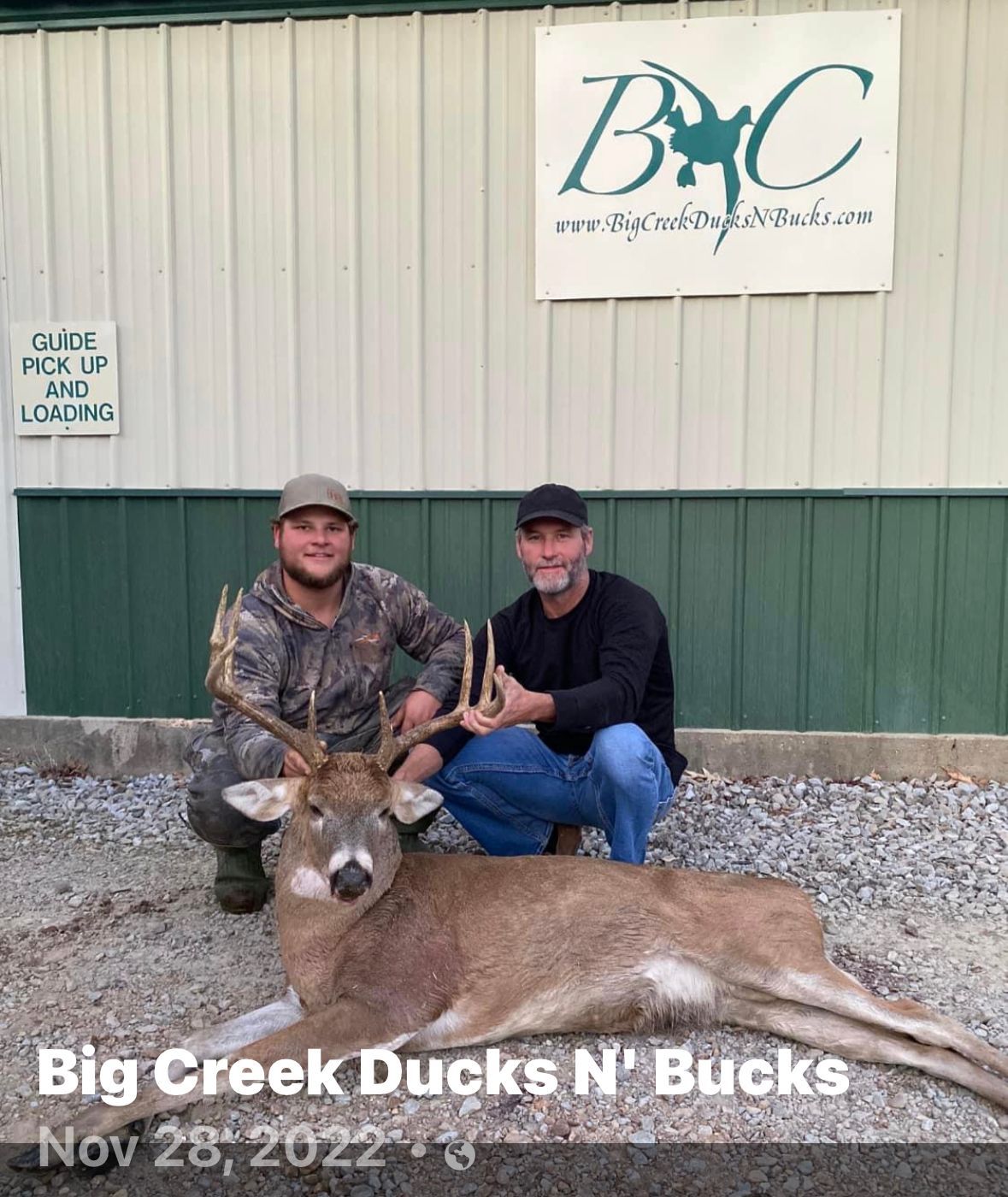 Two men kneel behind a deer trophy in front of a Big Creek Ducks N' Bucks building