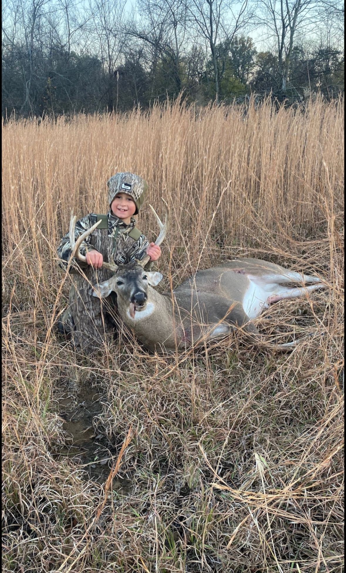 Hunter kneeling beside a deer in tall grass, holding up the animal’s antlers.