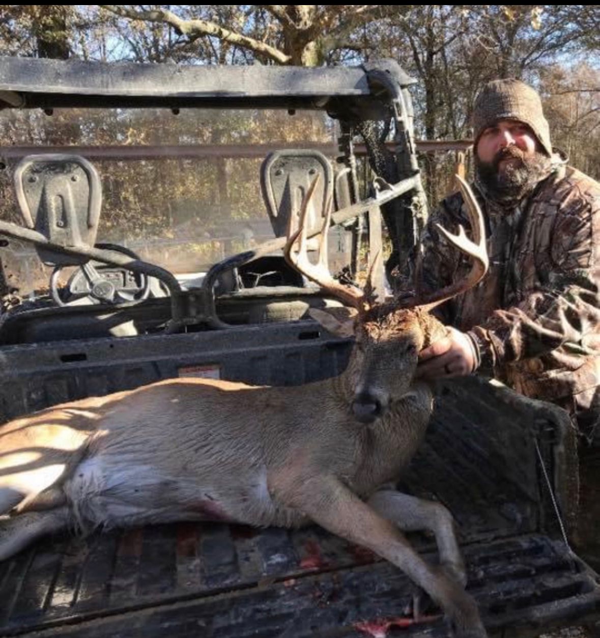 Hunter kneeling beside a deer in the back of a muddy off-road vehicle in a winter woods setting